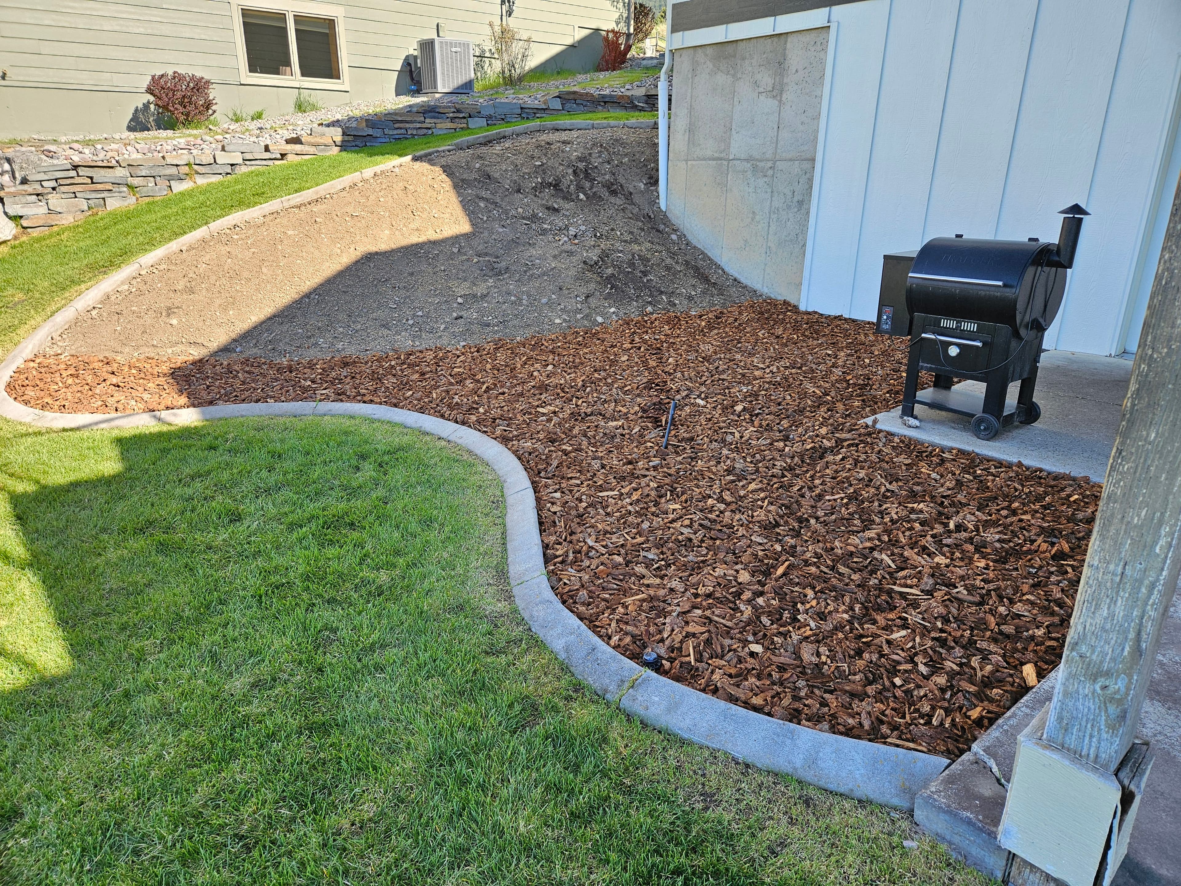 Landscaped backyard with mulch, curved stone border, and a smoker grill.