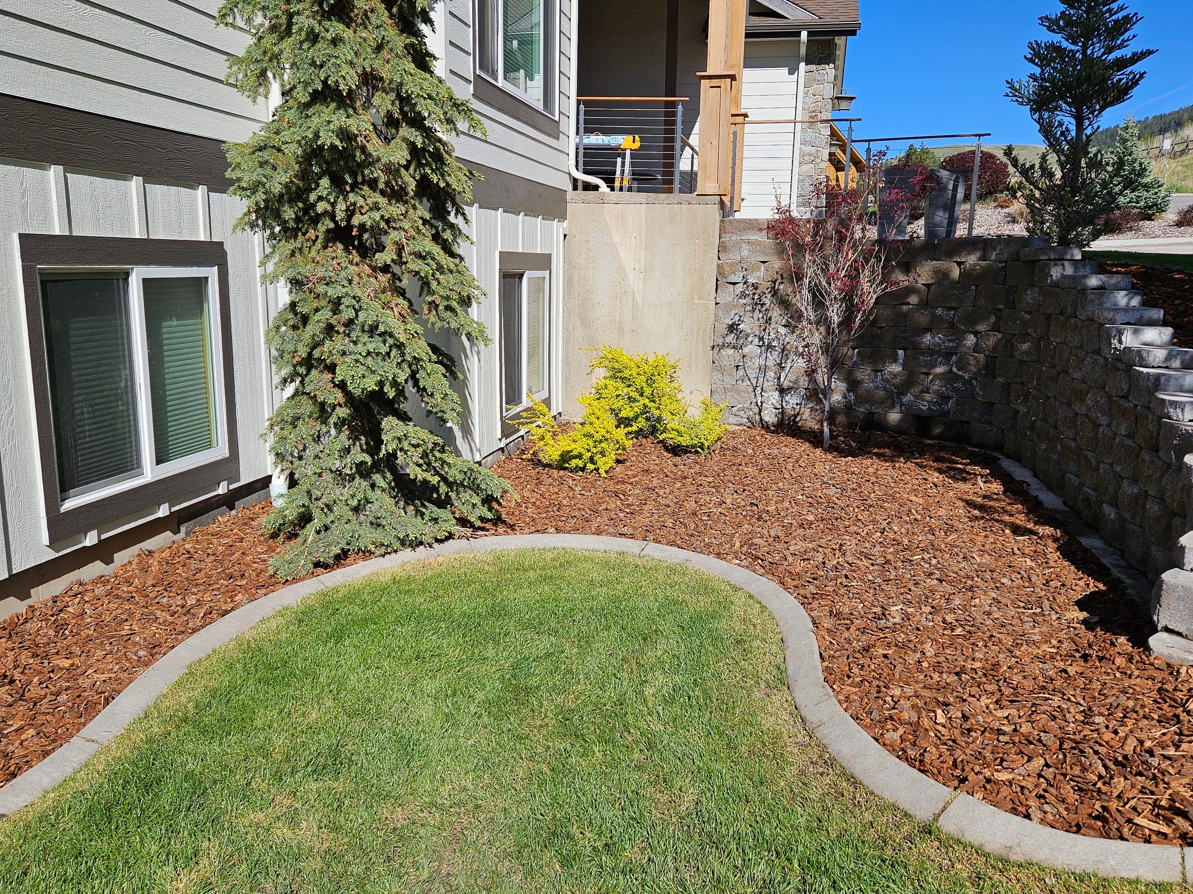 Landscaped home garden with green grass, shrubs, and decorative mulch beside a stone wall.