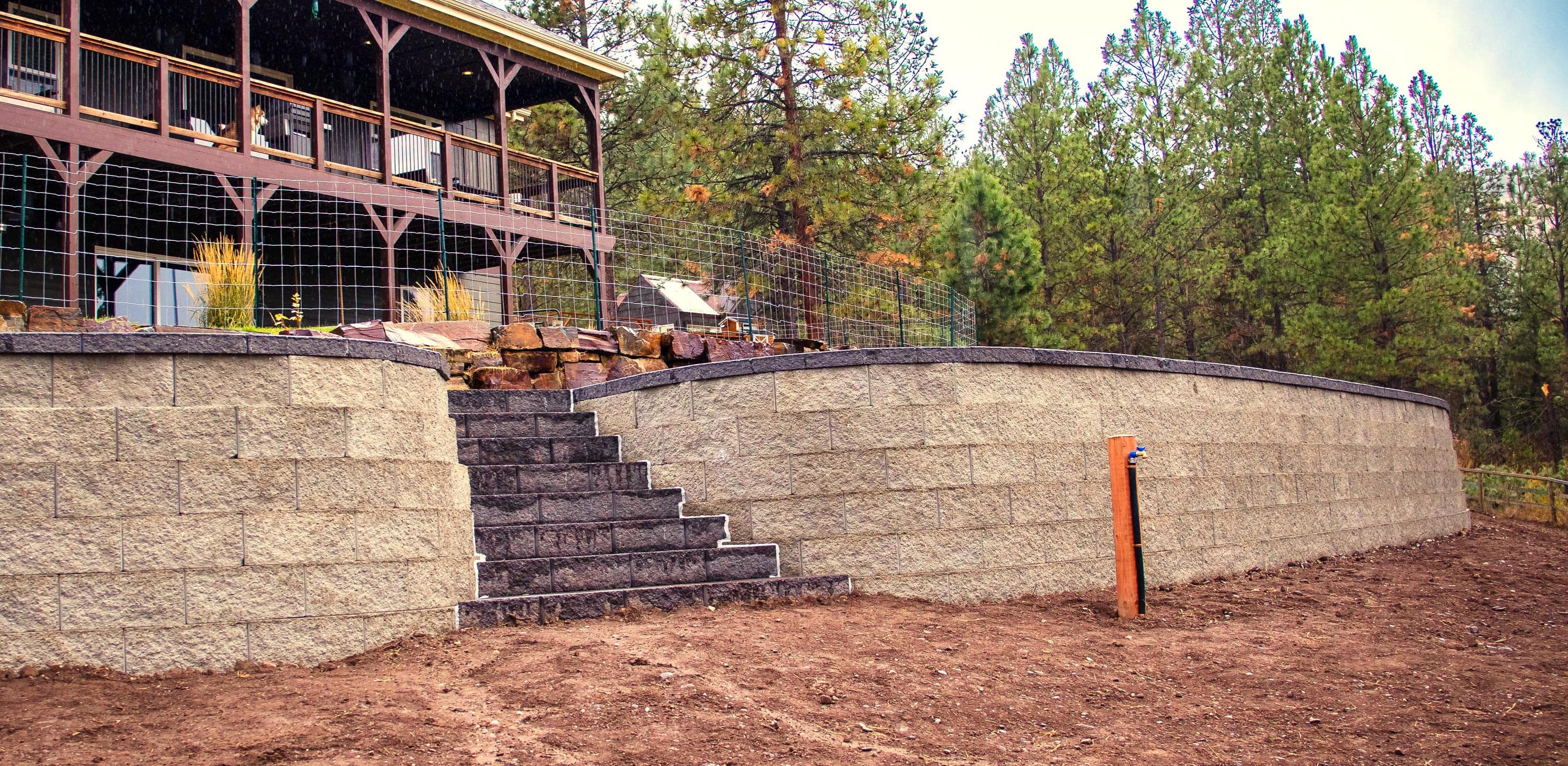 Modern stone retaining wall with stairs leading to a wooden deck surrounded by pine trees.