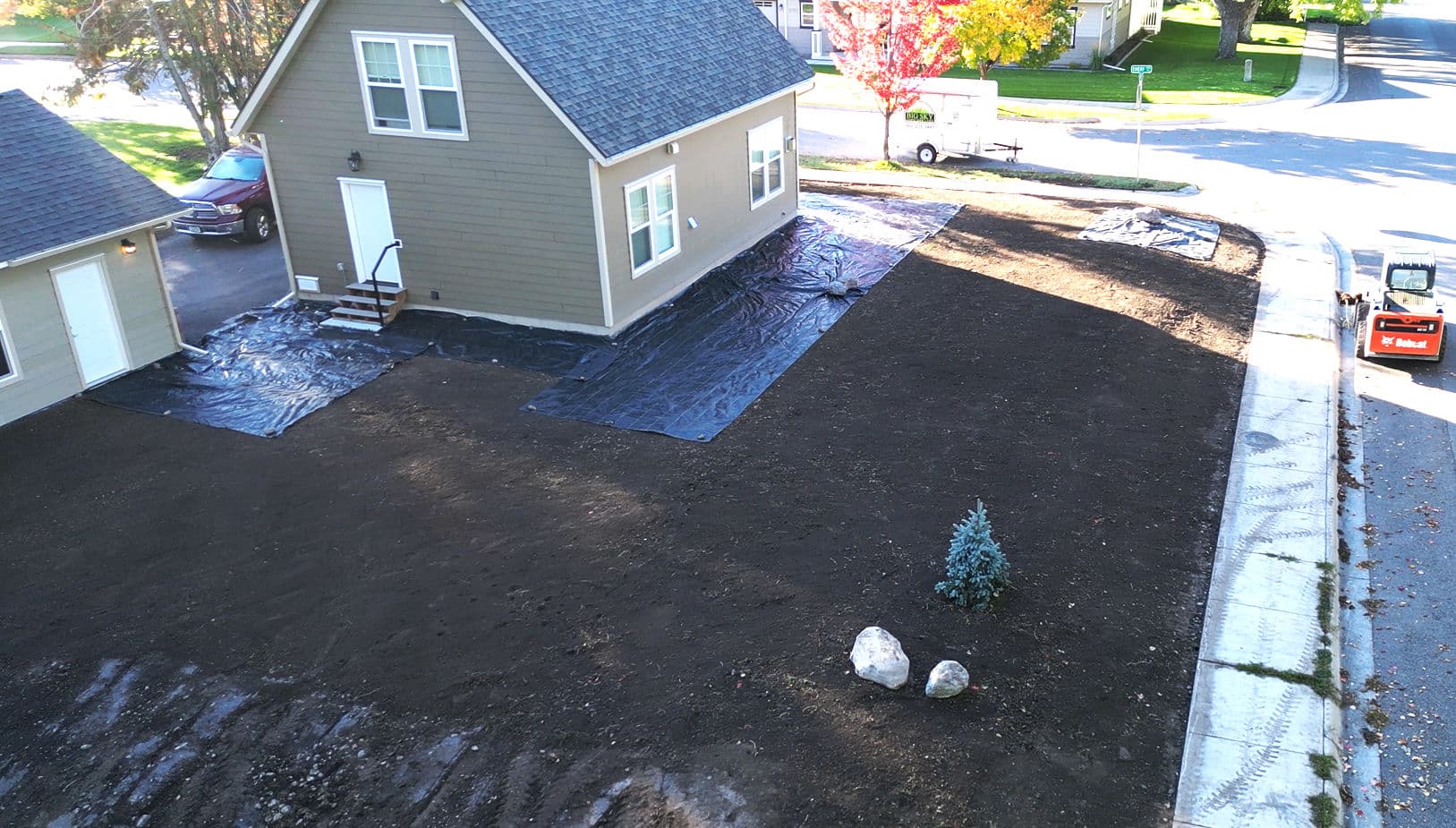 Aerial view of a residential property with freshly landscaped yard, black tarp, and small tree.