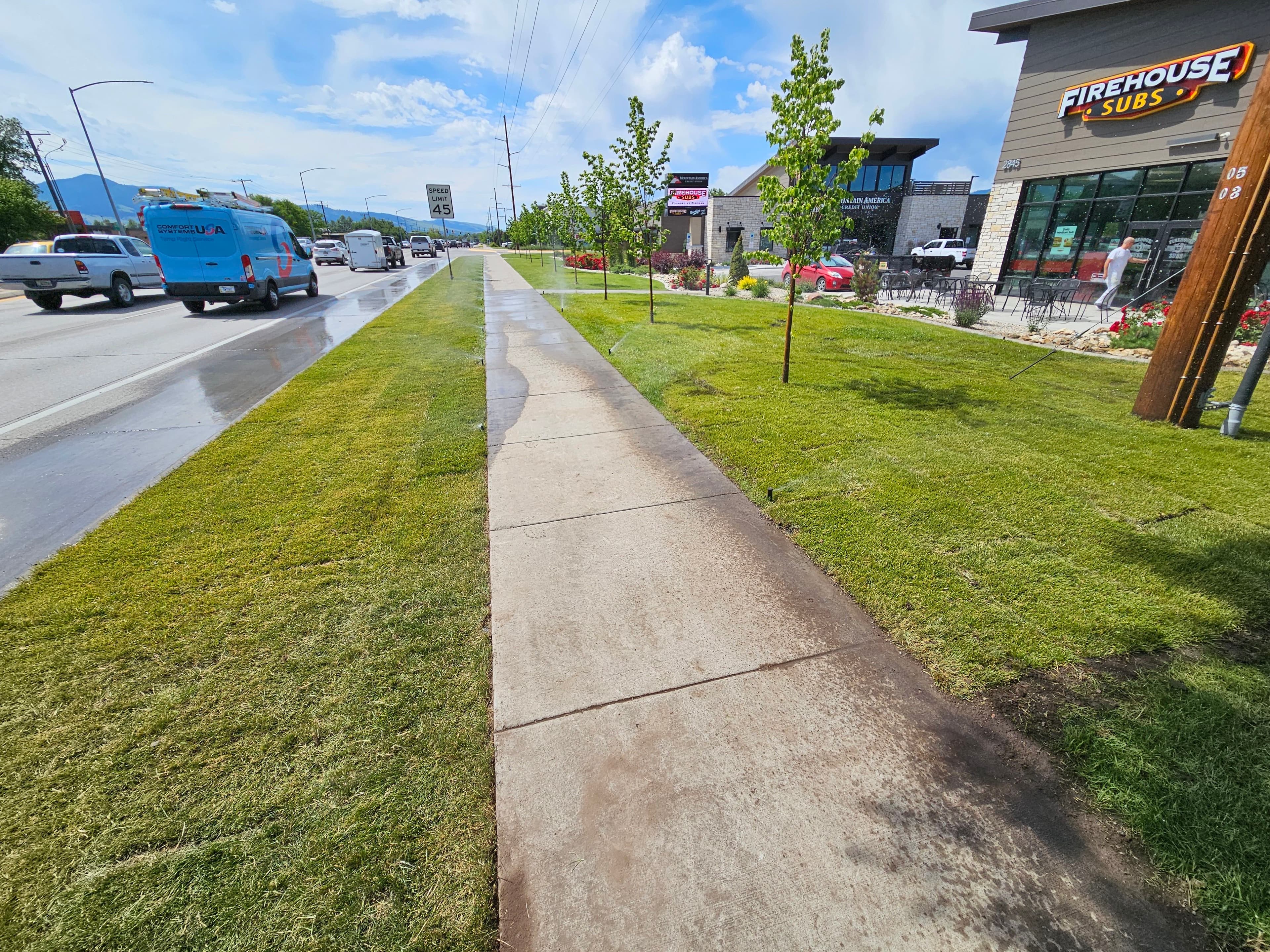 Sidewalk alongside a busy road with lush grass and a Firehouse Subs restaurant nearby.