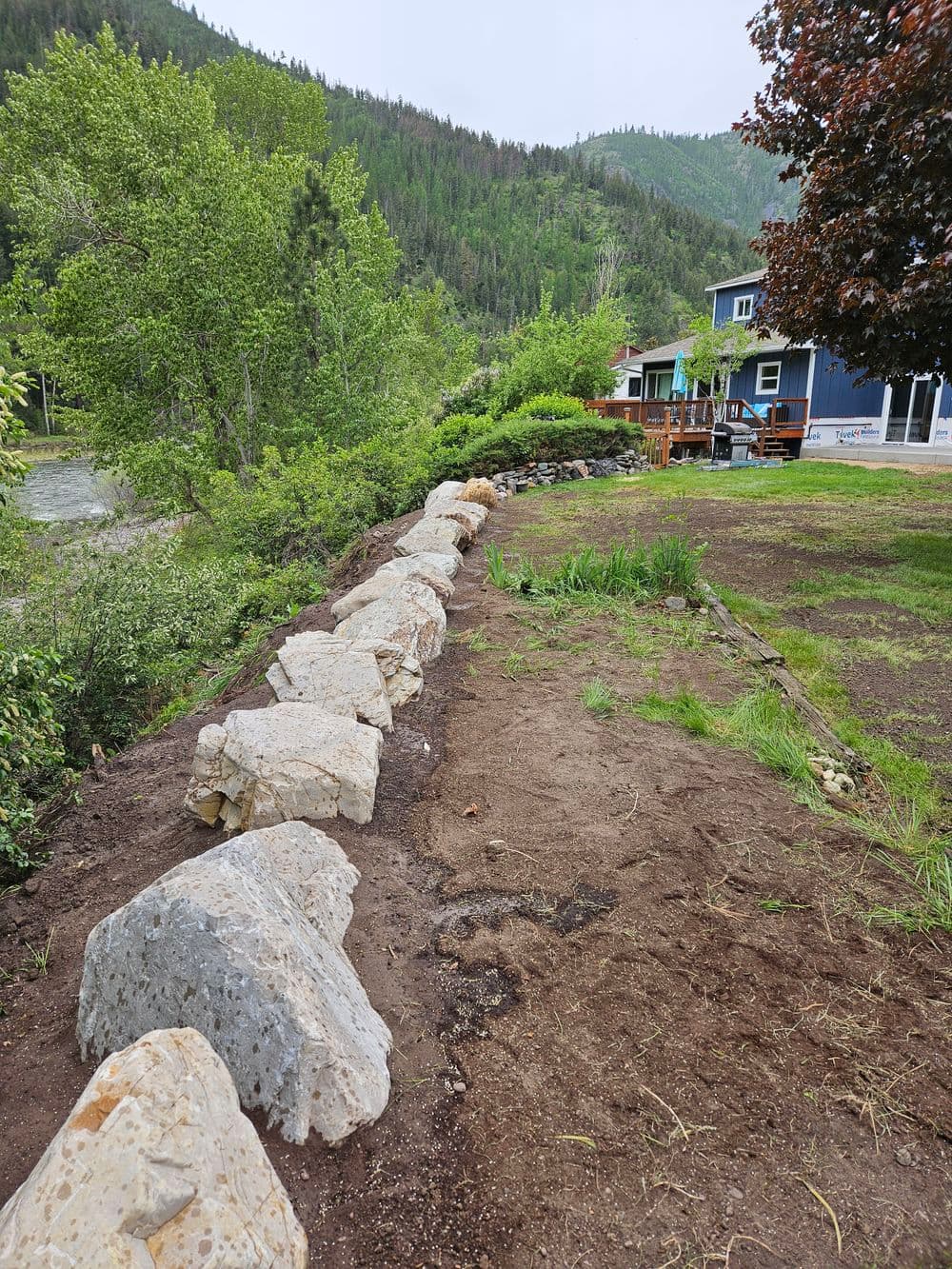 Rock retaining wall in a green landscape near a blue house and mountain backdrop.