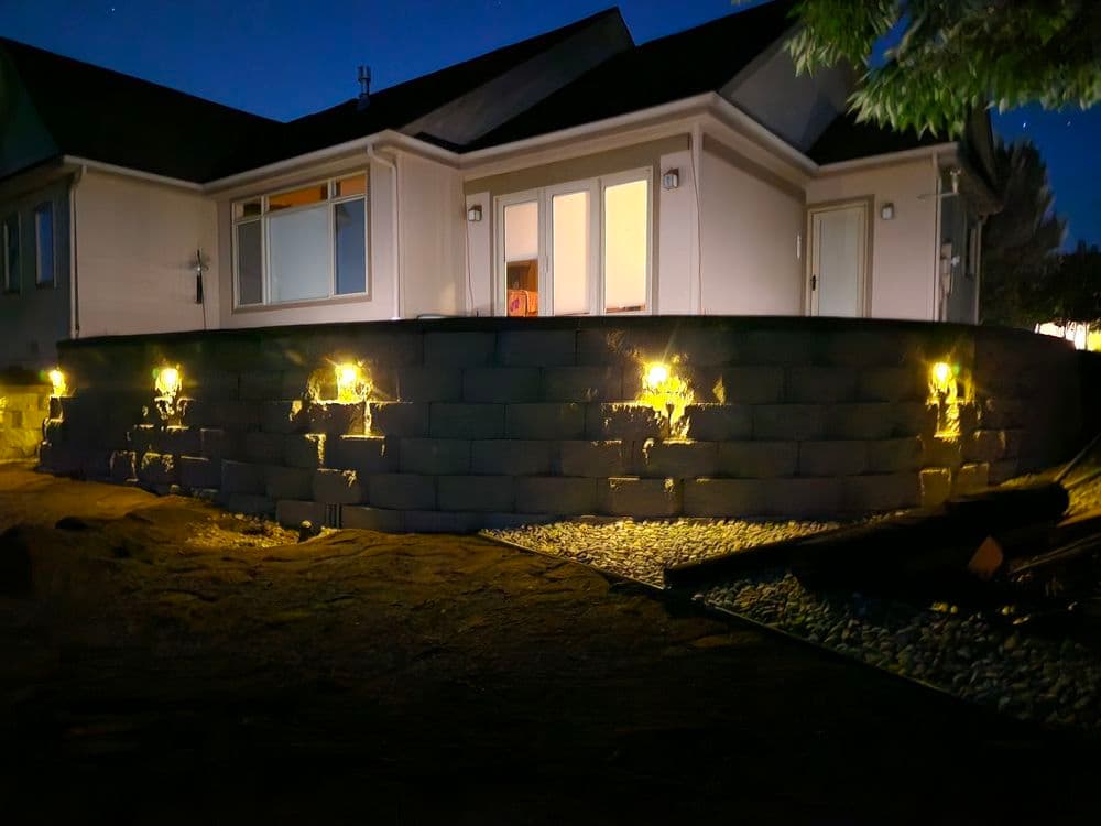Modern home exterior at night with illuminated stone wall landscaping and bright windows.